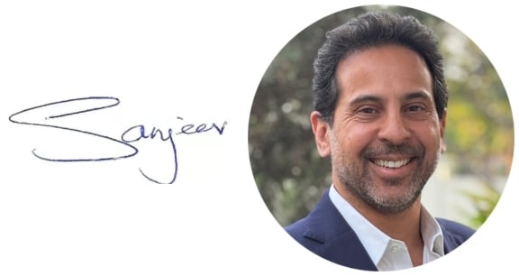 Professional headshot of a smiling man in a navy blazer and white shirt, this is the MD of Keylink Sanjeev,shown alongside a handwritten signature reading “Sanjeev”. The portrait is presented within a circular frame against a light grey background.